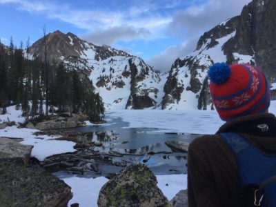 Man in a hat sits looking at a partially frozen lake surrounded by snowy cliffs.