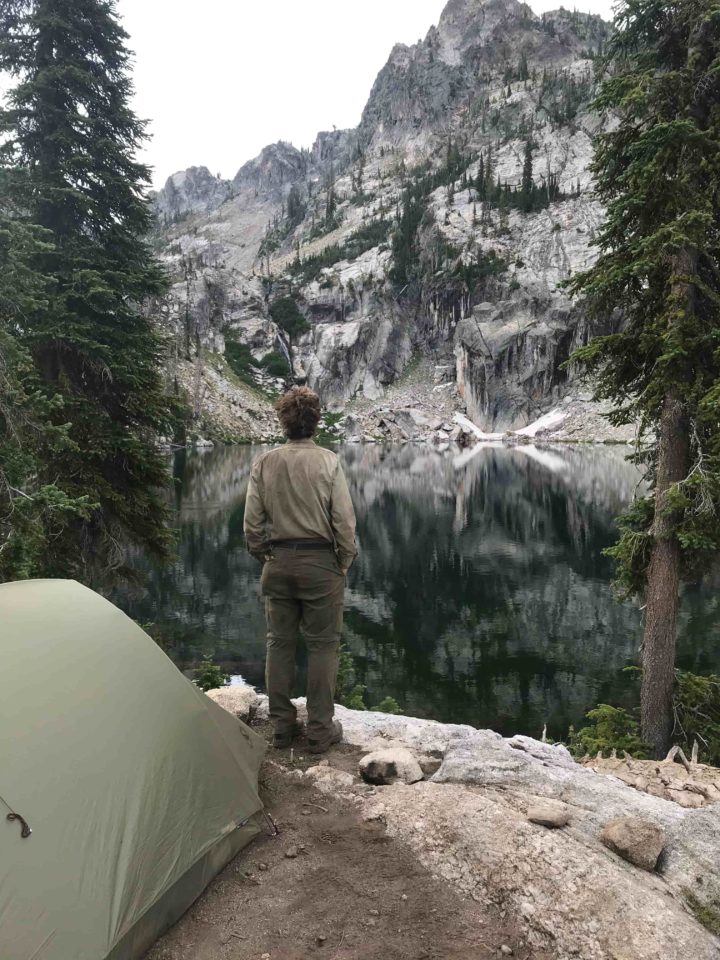 Wilderness ranger stands by his tent at trail creek lake with a jagged ridge and cliffs across the lake.