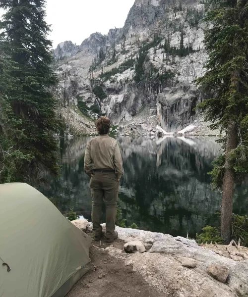 Wilderness ranger stands by his tent at trail creek lake with a jagged ridge and cliffs across the lake.